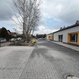 Budomad Michał Domagała - Nowoczesny budynek z szarymi ścianami i żółtymi obramowaniami okien, otoczony nową kostką brukową. Widoczny sprzęt budowlany i drzewo bez liści.