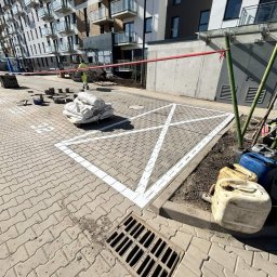 Budomad Michał Domagała - Oznakowane białą farbą miejsce parkingowe na kostce brukowej, z widocznym pracownikiem w tle i materiałami budowlanymi. Nowe budownictwo.