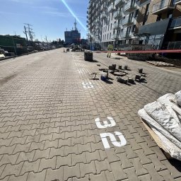 Budomad Michał Domagała - Układanie kostki brukowej na chodniku z numerem 32, w tle budynek mieszkalny i pracownik w żółtej kamizelce. Widoczne narzędzia i materiały budowlane.