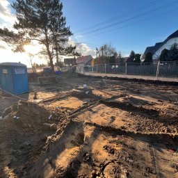 Budomad Michał Domagała - Przygotowanie terenu pod budowę domu: wykopane fundamenty, widoczna ziemia, tymczasowa toaleta budowlana, ogrodzenie i domy w tle, słoneczny dzień.