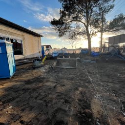 Budomad Michał Domagała - Budowa domu w toku: surowy budynek z oknami, przenośna toaleta, sprzęt budowlany i wykopana ziemia na działce w słoneczny dzień. Widoczne materiały budowlane.