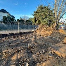 Budomad Michał Domagała - Przygotowanie terenu pod budowę: wykop z widocznymi śladami opon, ogrodzenie tymczasowe, w tle dom jednorodzinny i maszyna budowlana. Słoneczny dzień.