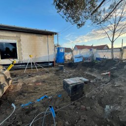 Budomad Michał Domagała - Fundamenty w trakcie budowy: wykop z widoczną studzienką, częściowo wymurowana ściana budynku z oknem, narzędzia pomiarowe i niebieski przenośny WC.
