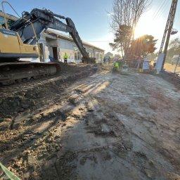 Budomad Michał Domagała - Żółta koparka Volvo na placu budowy domu z szarego bloczka. W tle pracownicy w kamizelkach odblaskowych i jasne słońce.