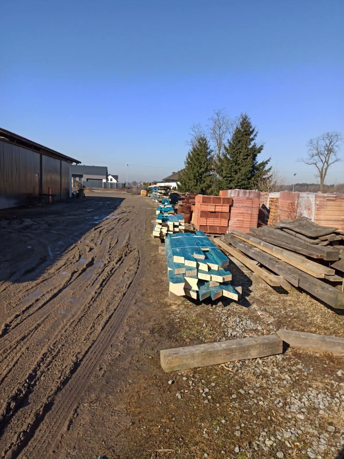 Składowane materiały budowlane: drewno impregnowane na niebiesko, cegła ceramiczna i drewniane belki na błotnistym placu budowy w słoneczny dzień.