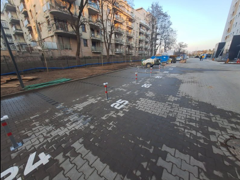 Oznakowane, mokre kostki brukowe na parkingu z numerami miejsc, w tle budynki mieszkalne i sprzęt budowlany. Widoczne słupki oddzielające miejsca parkingowe.