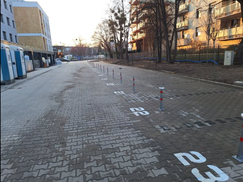 Ukończony parking z kostki brukowej z numeracją miejsc i słupkami zabezpieczającymi, widok z perspektywy wjazdu, budynki w tle.