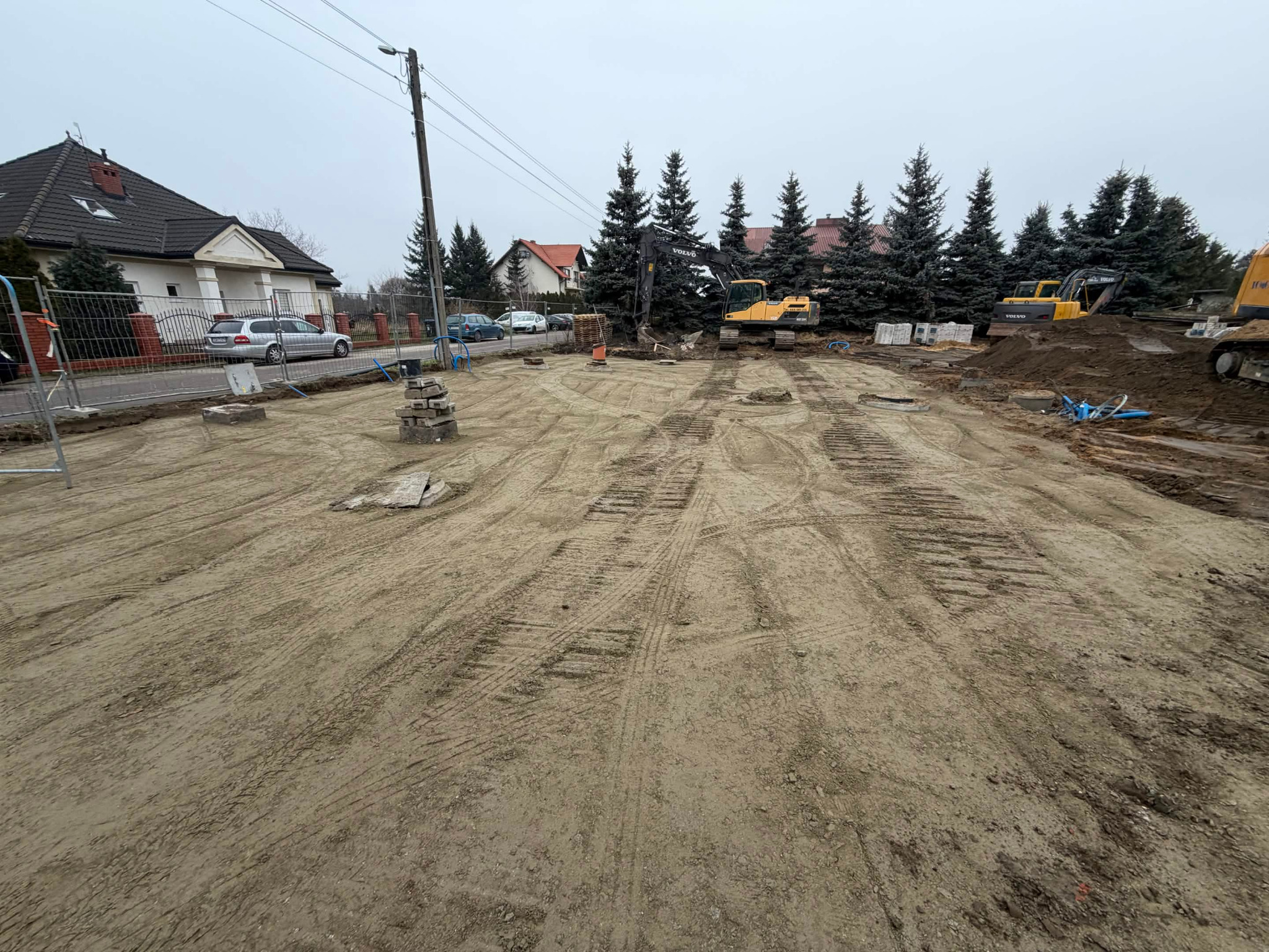 Przygotowanie terenu pod budowę domu: wyrównana działka z widocznymi śladami opon koparek, w tle domy jednorodzinne i wysokie drzewa iglaste.