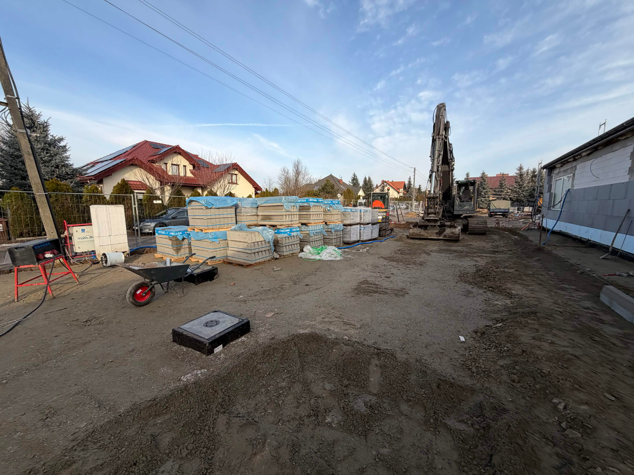 Plac budowy z paletami bloczków, koparką i częściowo wymurowaną ścianą budynku w tle, w słoneczny dzień. Widoczna taczka i inne narzędzia budowlane.