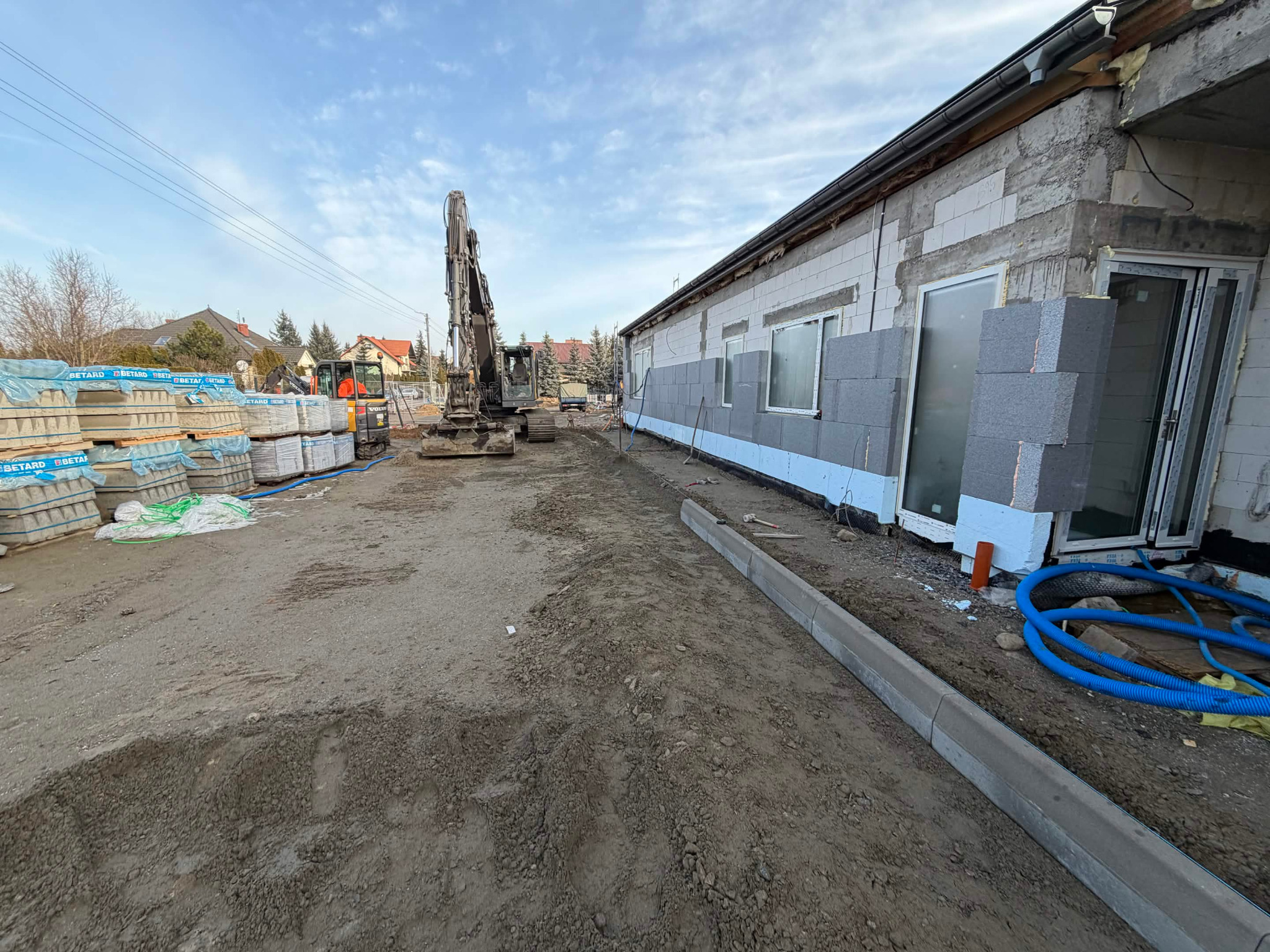 Budowa domu: ściany z betonu komórkowego, izolacja, okna. Na placu budowy palety z bloczkami i koparka. Widok na budynek w trakcie wznoszenia.