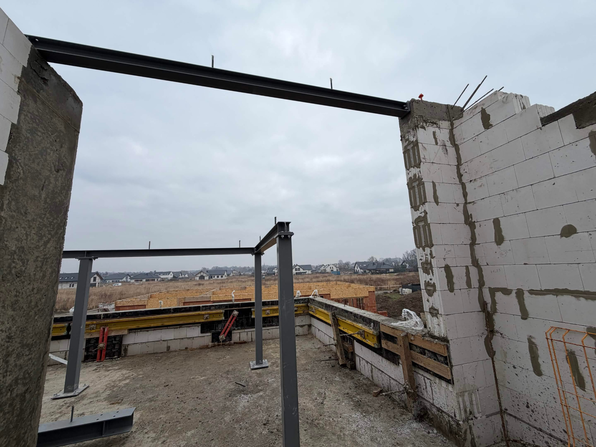 Wznoszenie ścian z bloczków betonu komórkowego, wzmocnionych stalowymi belkami. Widoczne szalunki fundamentów i konstrukcja nośna. Budowa domu w trakcie realizacji.