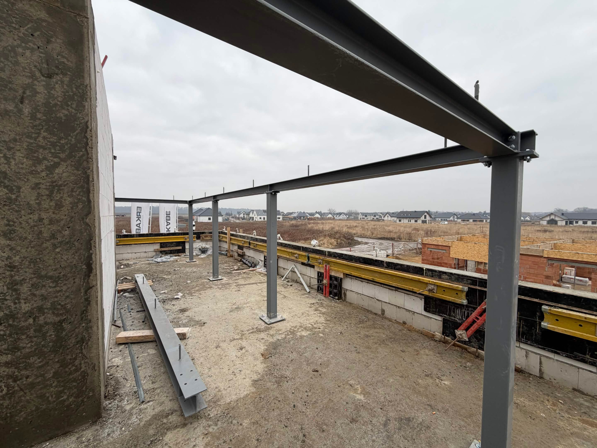 Konstrukcja stalowa z kolumnami na fundamencie domu w budowie, widok na okoliczne domy i pochmurne niebo. Elementy szalunków widoczne w tle.