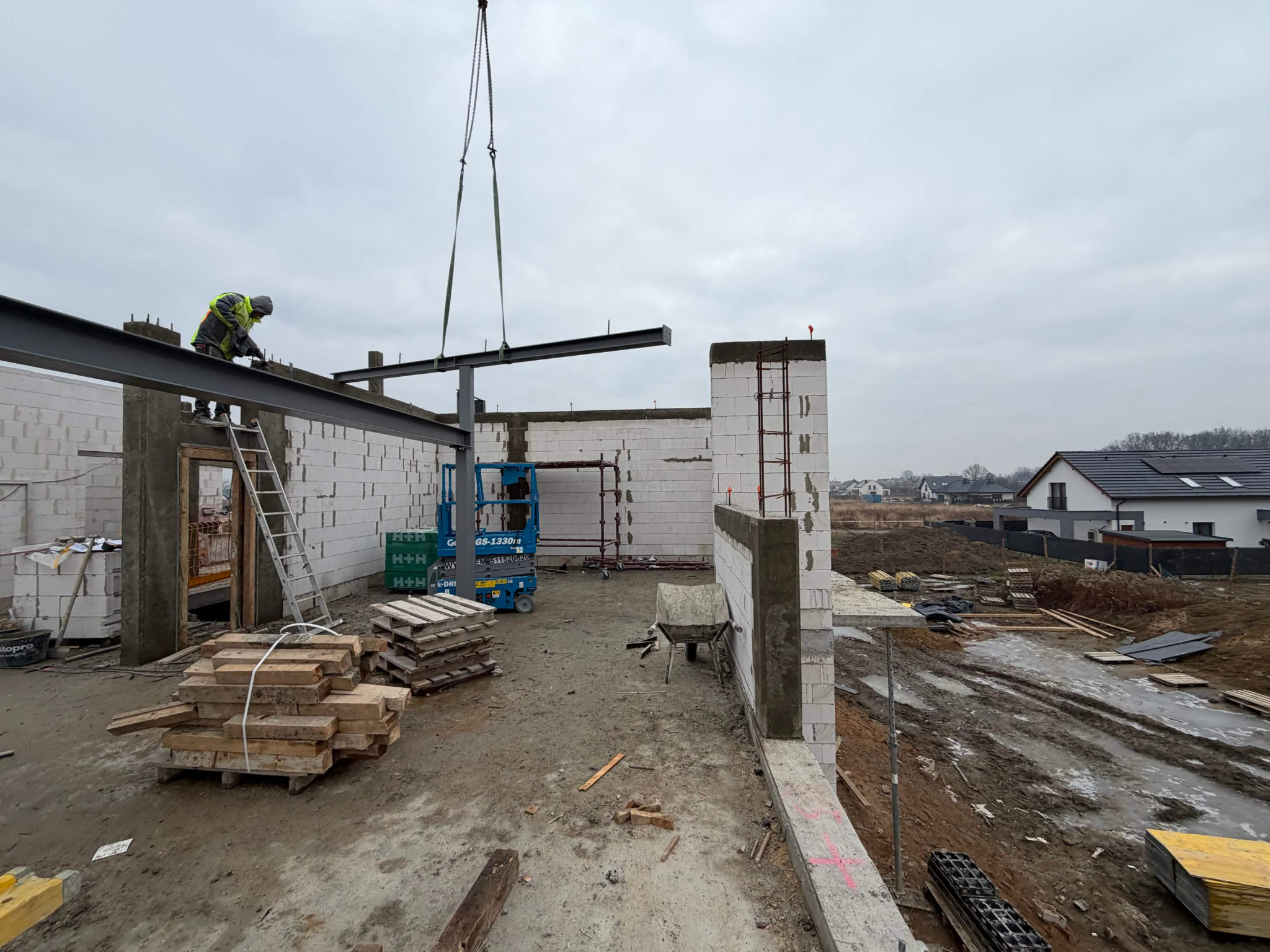 Montaż stalowej belki dźwigiem na budowie domu z białych bloczków. Widoczny pracownik na drabinie, podnośnik i palety z materiałami. Tło: zabudowa jednorodzinna.