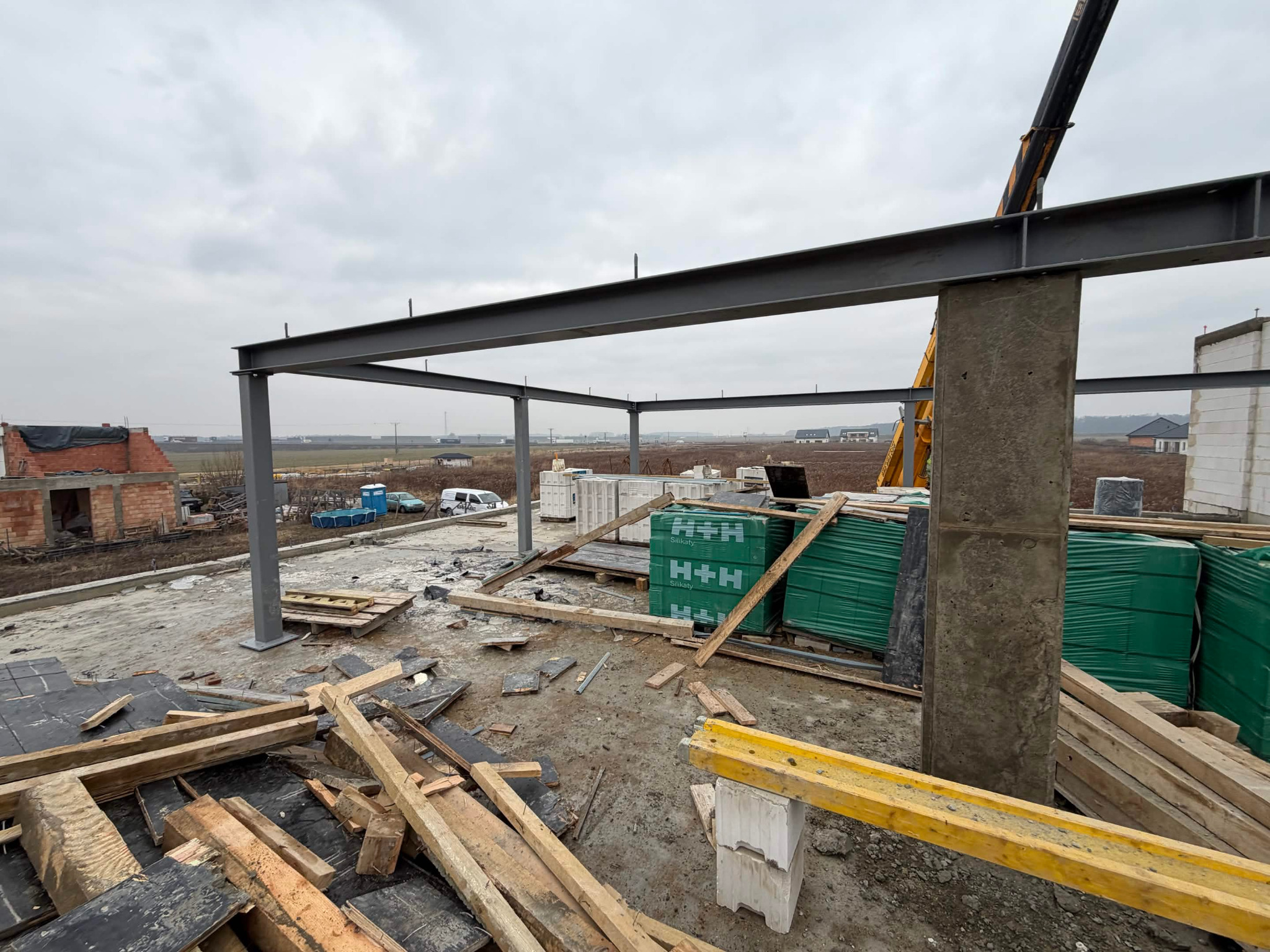 Konstrukcja domu w trakcie budowy: stalowy szkielet, bloczki silikatowe H+H, drewniane elementy szalunkowe, dźwig w tle, widok na okolicę. Etap wznoszenia.
