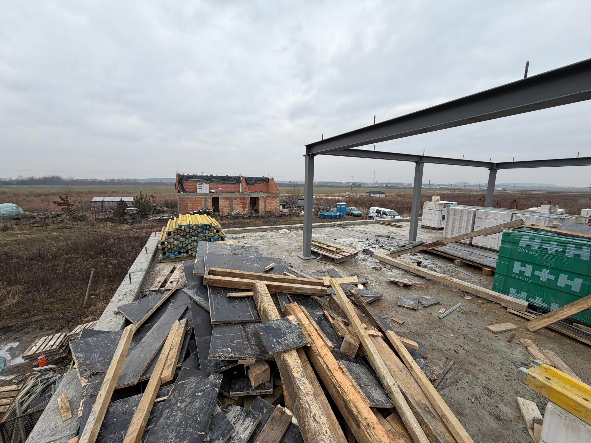 Budowa domu w trakcie: stalowa konstrukcja, palety z bloczkami silikatowymi H+H, drewniane deski i w tle budynek w stanie surowym otwartym, pochmurne niebo.