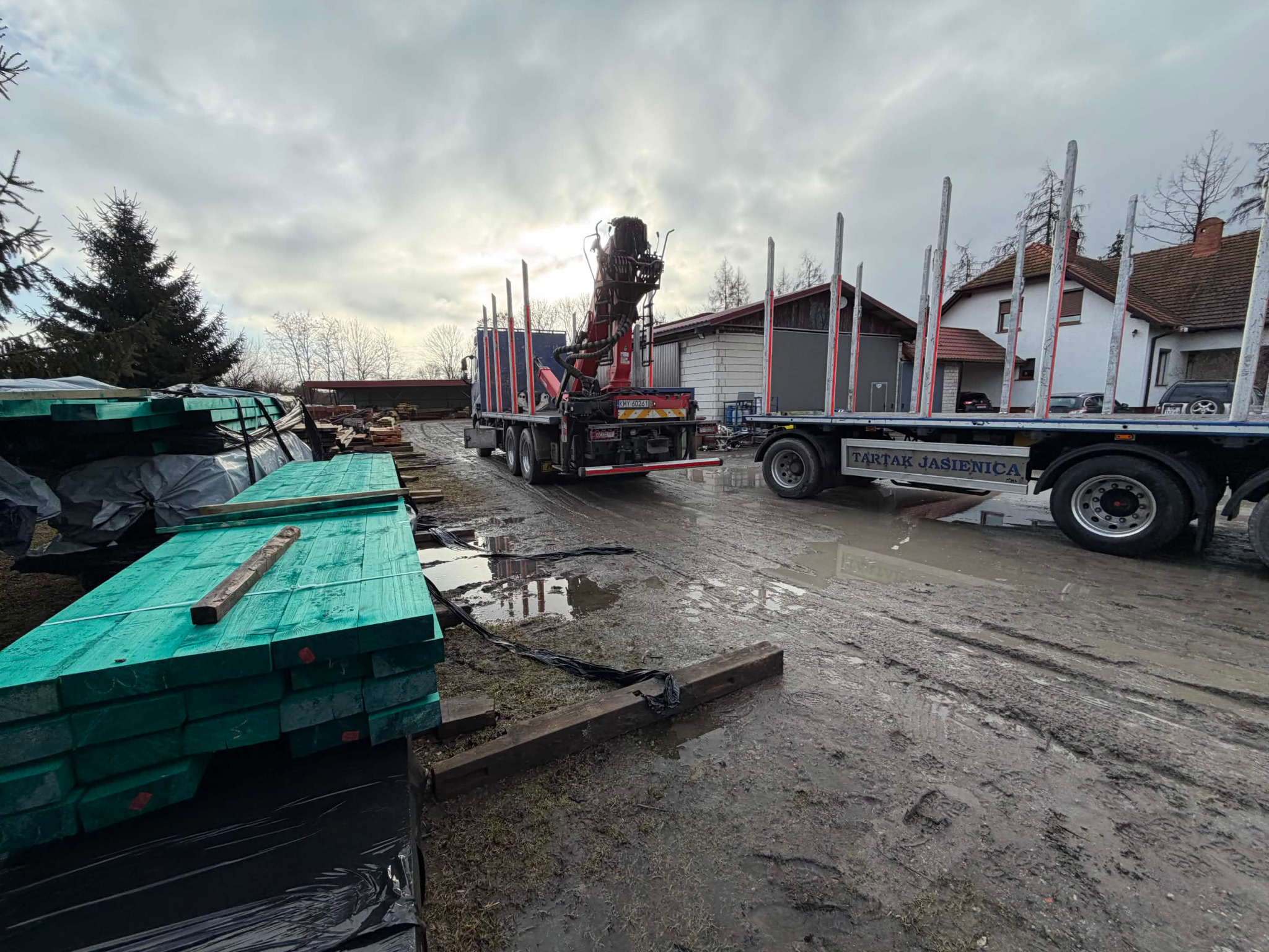 Ciężarówka z dźwigiem i przyczepą Tartak Jasienica na błotnistym terenie, obok stos turkusowych desek. Pochmurne niebo w tle.