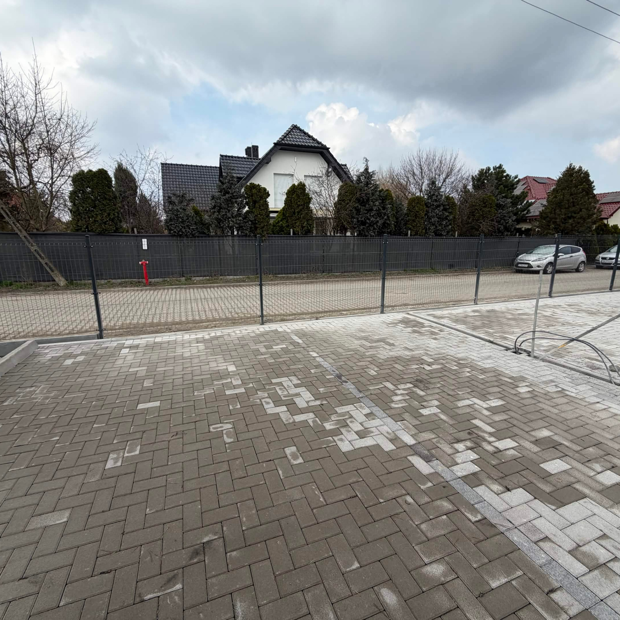 Układanie kostki brukowej na podjeździe domu jednorodzinnego. Widoczne fragmenty niezakończonej nawierzchni i ogrodzenie posesji z czerwoną hydrantem.