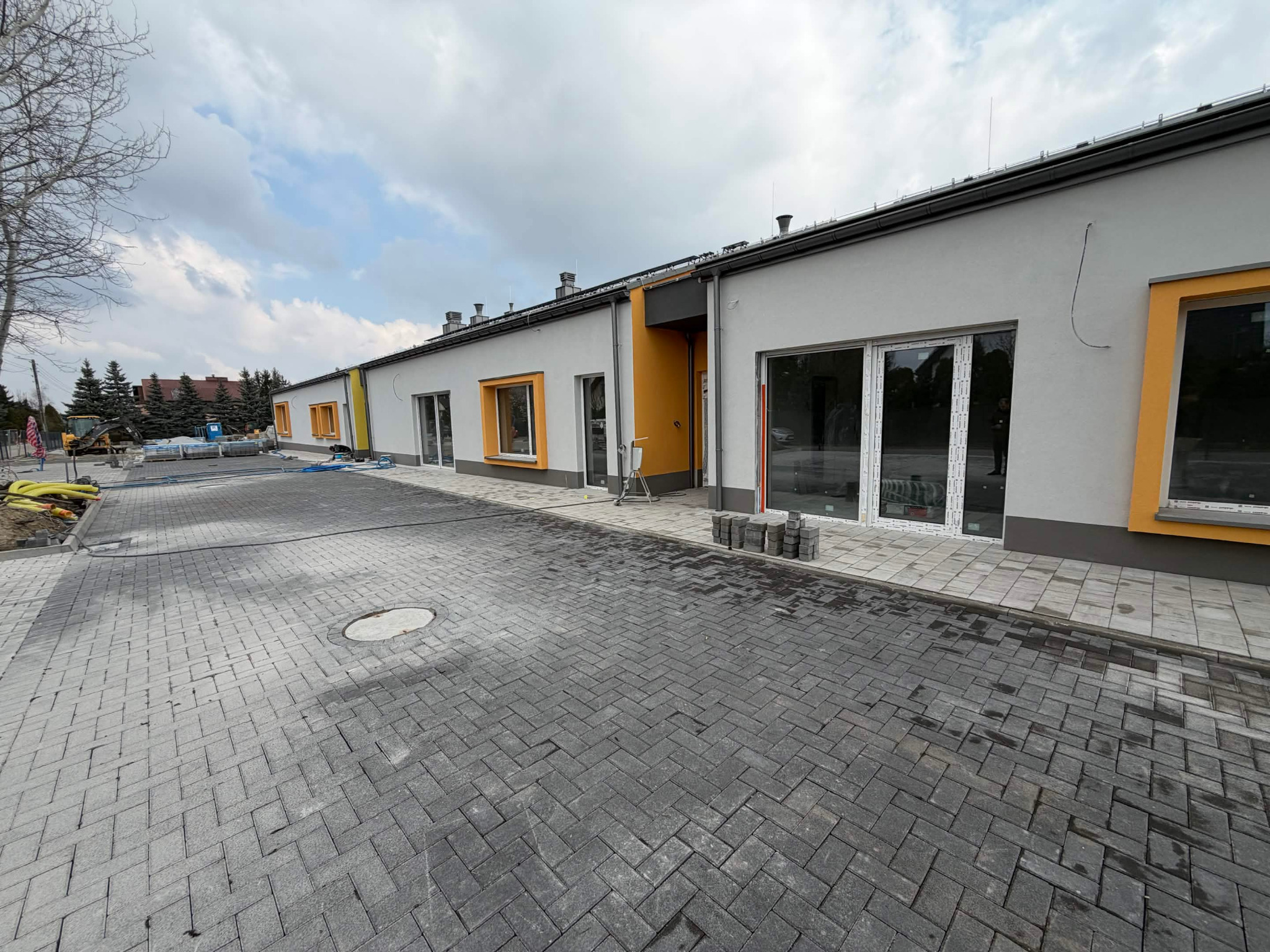 Parterowy budynek z szarymi ścianami i żółtymi akcentami okiennymi, widoczny z zewnątrz, z nową kostką brukową na podjeździe i elementami wykończeniowymi.