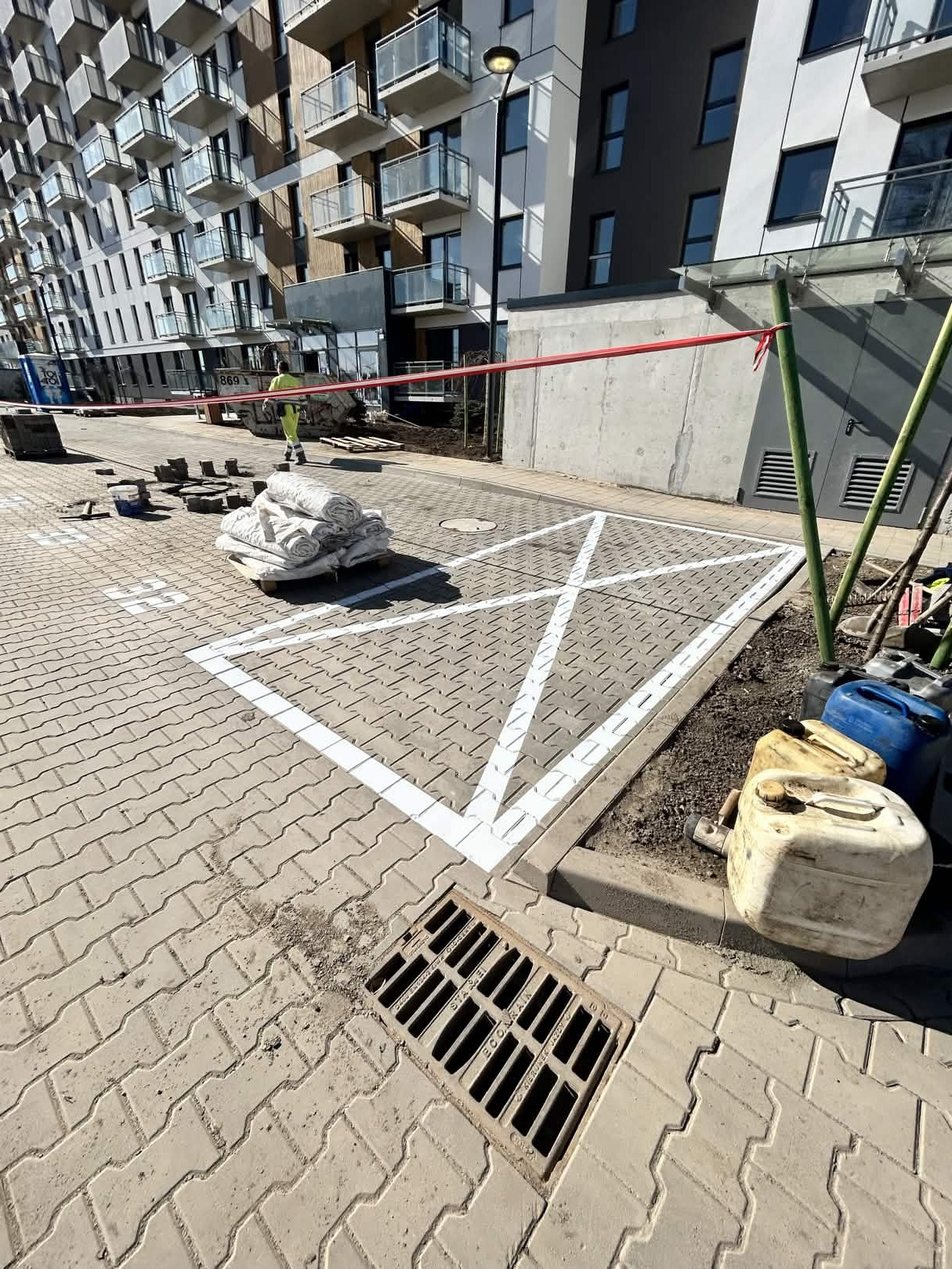 Oznakowane białą farbą miejsce parkingowe na kostce brukowej, z widocznym pracownikiem w tle i materiałami budowlanymi. Nowe budownictwo.