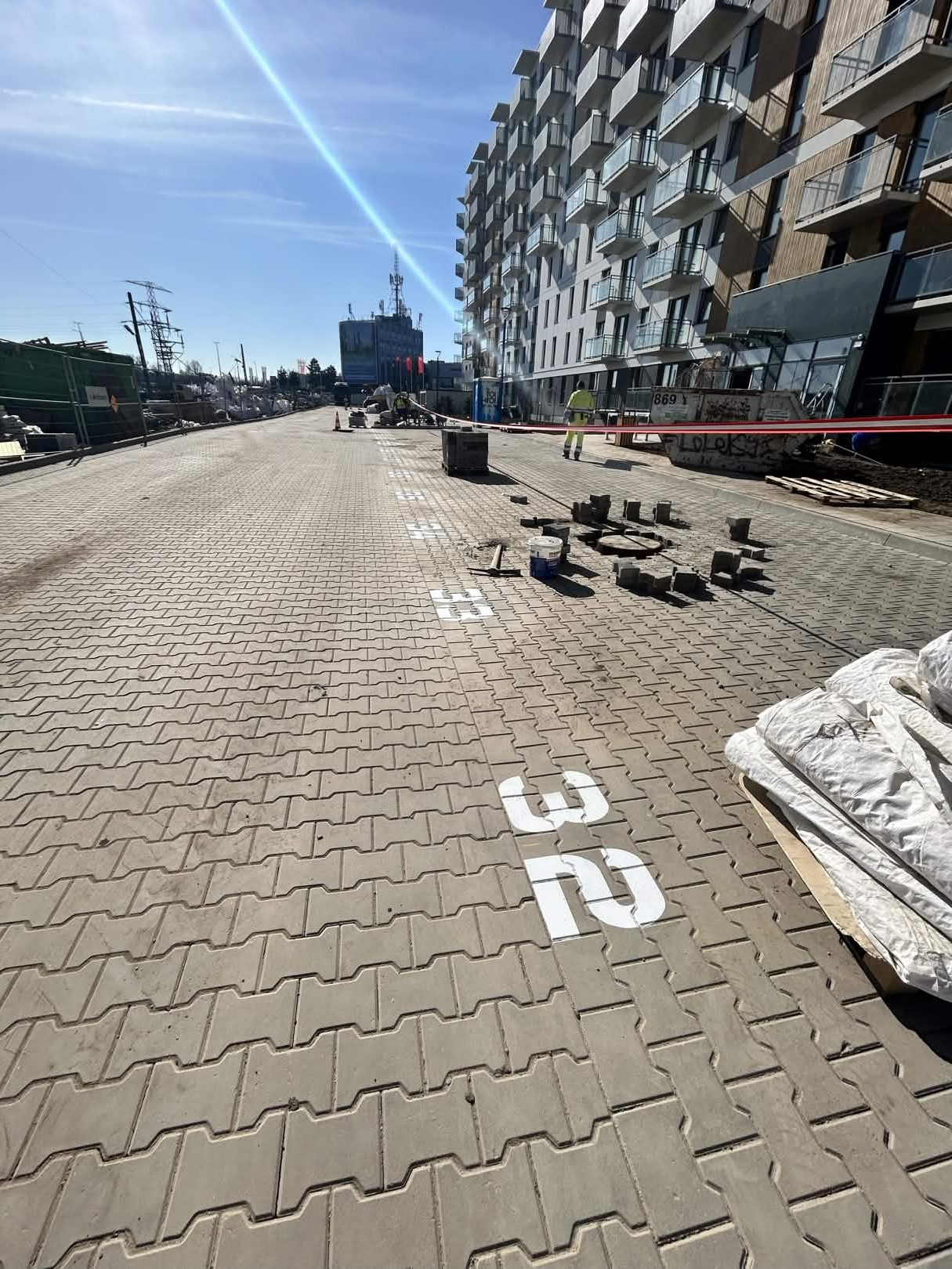 Układanie kostki brukowej na chodniku z numerem 32, w tle budynek mieszkalny i pracownik w żółtej kamizelce. Widoczne narzędzia i materiały budowlane.