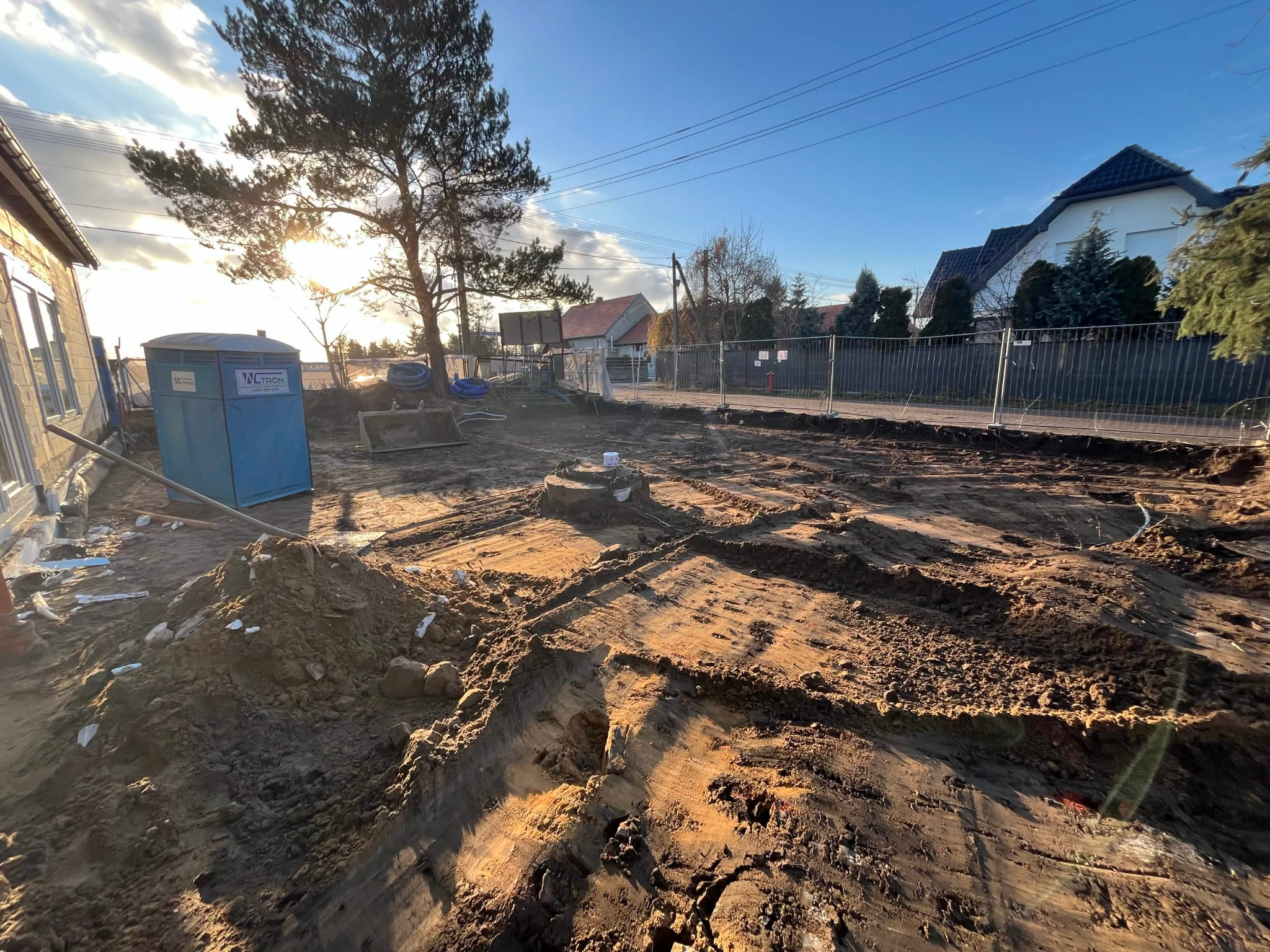 Przygotowanie terenu pod budowę domu: wykopane fundamenty, widoczna ziemia, tymczasowa toaleta budowlana, ogrodzenie i domy w tle, słoneczny dzień.