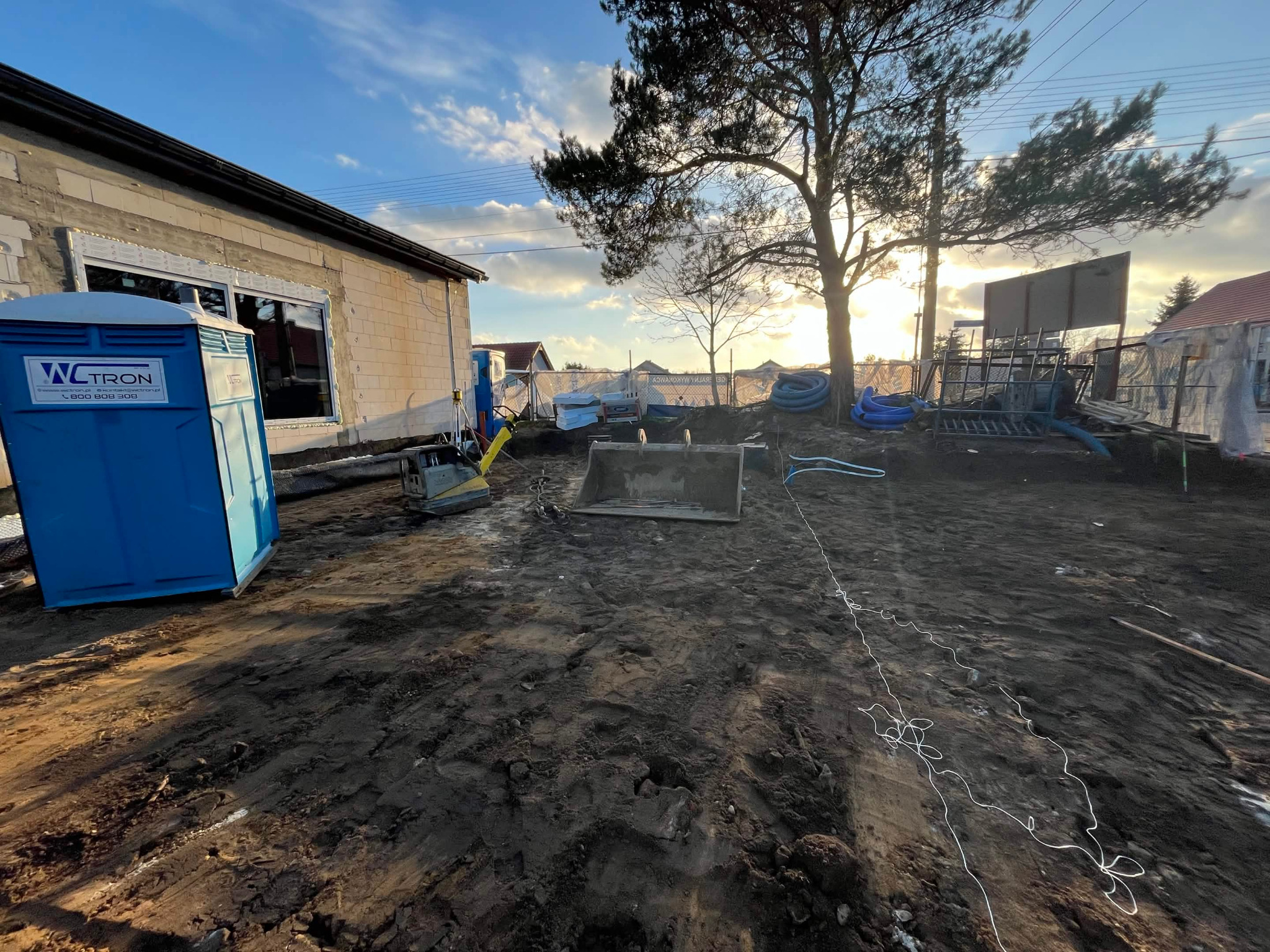 Budowa domu w toku: surowy budynek z oknami, przenośna toaleta, sprzęt budowlany i wykopana ziemia na działce w słoneczny dzień. Widoczne materiały budowlane.