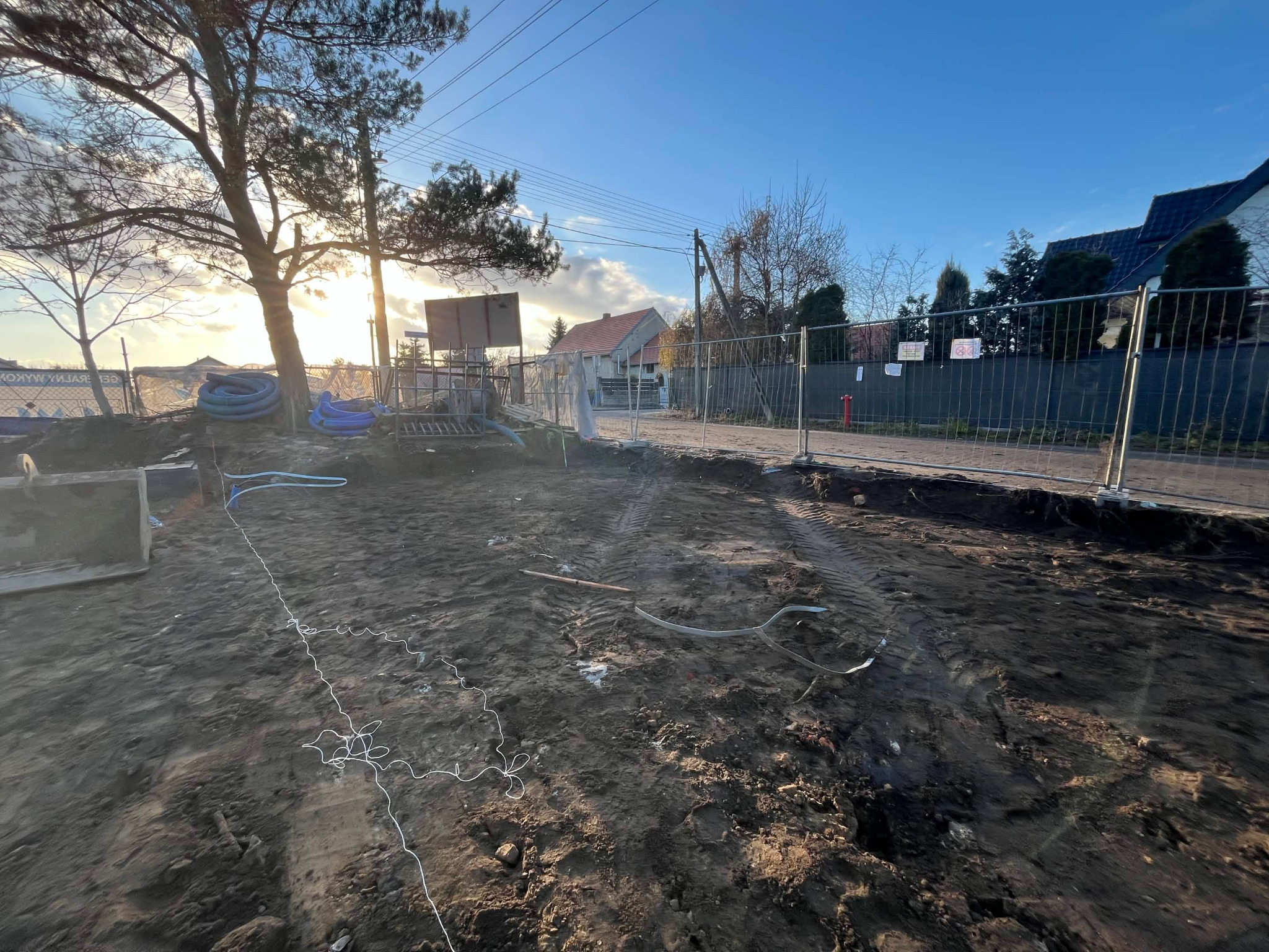 Teren budowy w przygotowaniu: wykopana ziemia, widoczne rury, ogrodzenie tymczasowe, w tle dom z czerwonym dachem i drzewa w promieniach zachodzącego słońca.