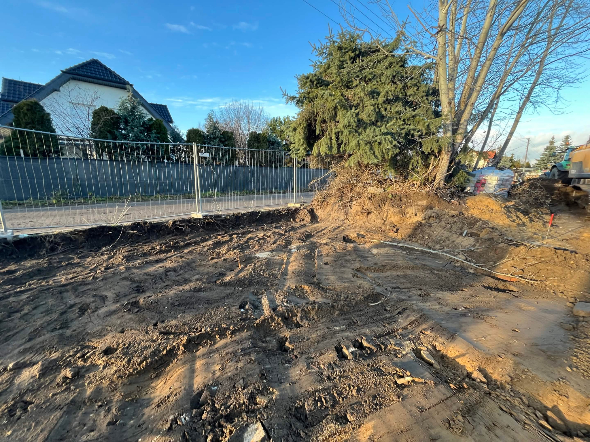 Przygotowanie terenu pod budowę: wykop z widocznymi śladami opon, ogrodzenie tymczasowe, w tle dom jednorodzinny i maszyna budowlana. Słoneczny dzień.