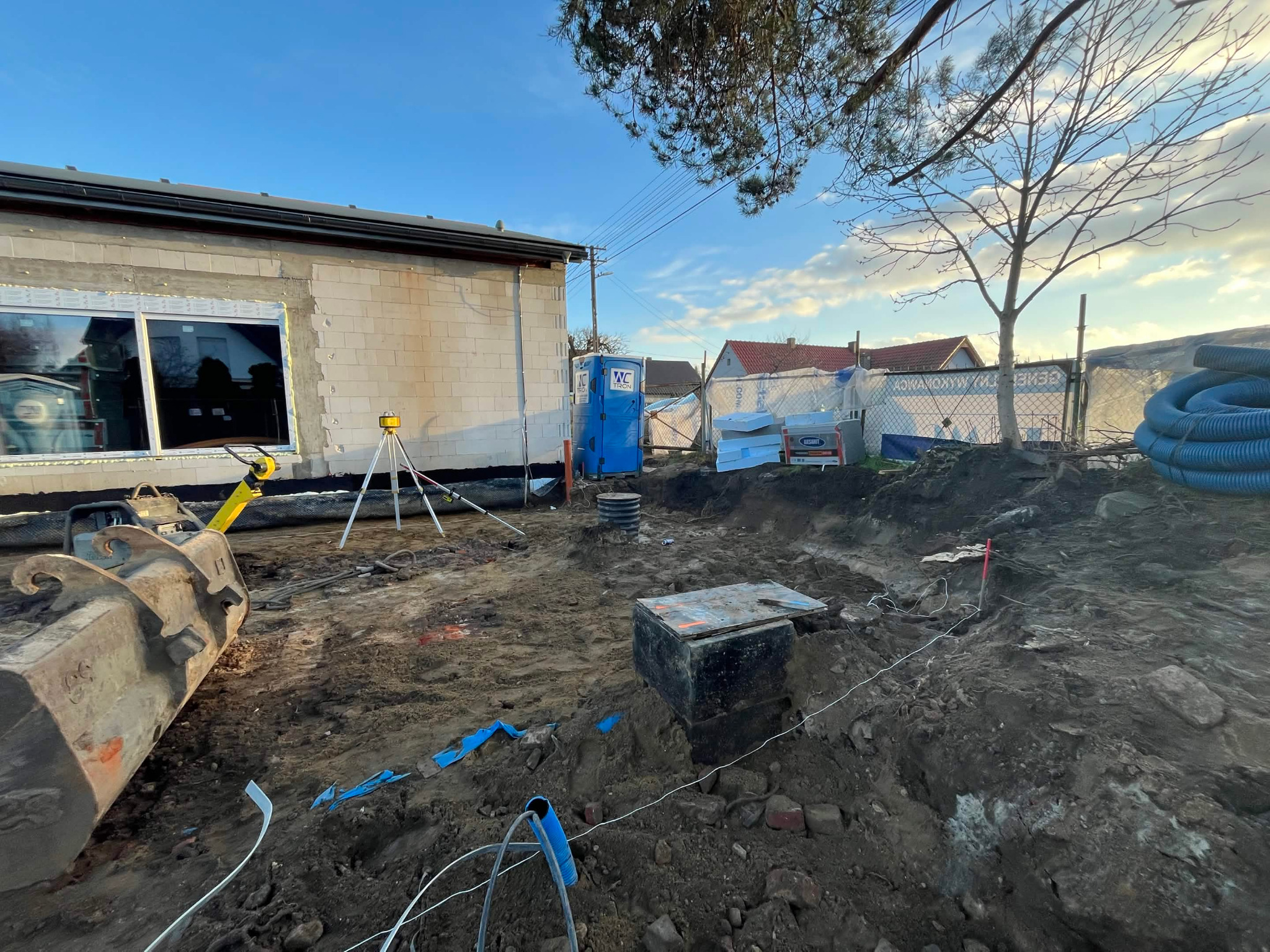 Fundamenty w trakcie budowy: wykop z widoczną studzienką, częściowo wymurowana ściana budynku z oknem, narzędzia pomiarowe i niebieski przenośny WC.