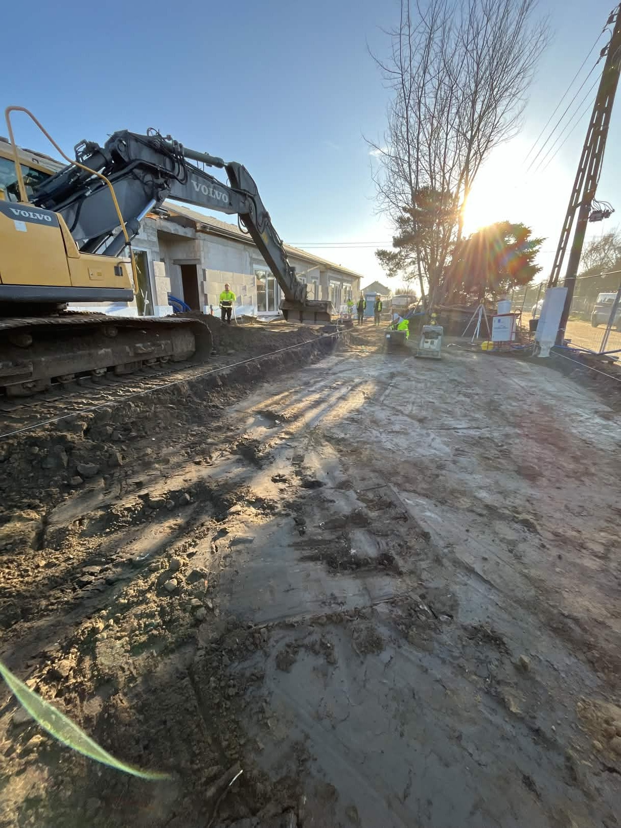 Żółta koparka Volvo na placu budowy domu z szarego bloczka. W tle pracownicy w kamizelkach odblaskowych i jasne słońce.