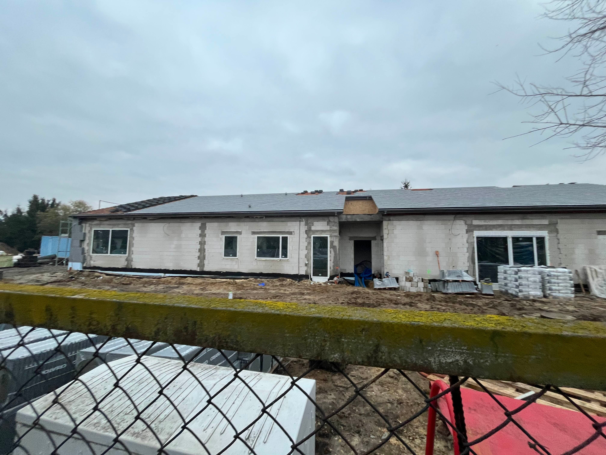 Budowa domu jednorodzinnego z szarej cegły, częściowo pokryty dachówką, widok zza ogrodzenia. Na pierwszym planie materiały budowlane.