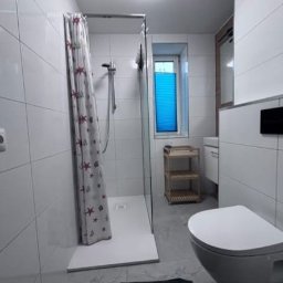 Patryk Kłosek - Biała łazienka po remoncie z kabiną prysznicową, zasłoną w gwiazdki, toaletą i oknem z niebieską roletą. Widok na wykończone wnętrze z płytkami.