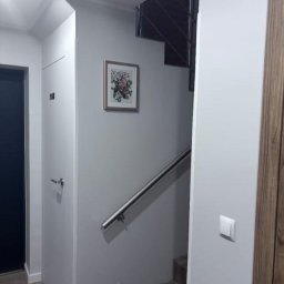 Patryk Kłosek - Szare ściany klatki schodowej z metalową poręczą, obrazkiem w ramce i fragmentem drewnianych drzwi. Widoczne schody i gniazdko elektryczne.