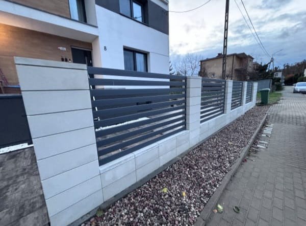 Nowoczesne ogrodzenie posesji z poziomymi, grafitowymi przęsłami i jasnymi, betonowymi słupkami. Wzdłuż ogrodzenia wysypany ozdobny żwir.
