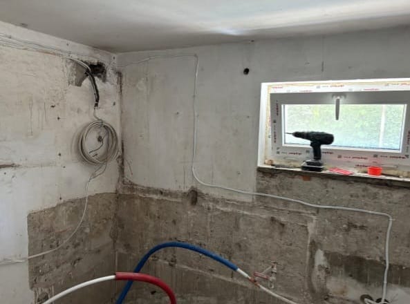 Surowe ściany w trakcie remontu z widocznymi przewodami elektrycznymi, rurami i wiertarką na parapecie okna. Etap prac przygotowawczych przed wykończeniem.