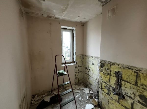 Wnętrze łazienki w trakcie demontażu starych płytek, widoczna drabina, ślady po kleju i resztki gruzu na podłodze. Okno z widokiem na zewnątrz.
