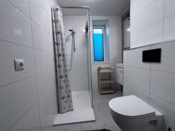 Biała łazienka po remoncie z kabiną prysznicową, zasłoną w gwiazdki, toaletą i oknem z niebieską roletą. Widok na wykończone wnętrze z płytkami.