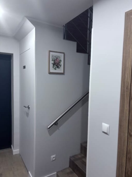 Szare ściany klatki schodowej z metalową poręczą, obrazkiem w ramce i fragmentem drewnianych drzwi. Widoczne schody i gniazdko elektryczne.