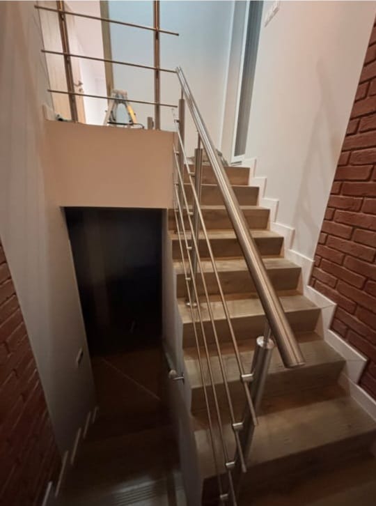 Schody z drewnianymi stopniami i metalową balustradą, ściana wykończona cegłą, widoczne wejście do pomieszczenia pod schodami. Oświetlenie górne.