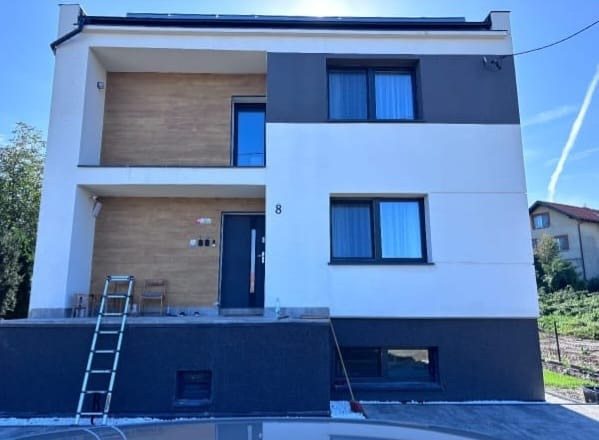 Dwukondygnacyjny dom po remoncie z numerem 8, z szarą elewacją na dole i białymi ścianami na górze. Widoczna drabina i częściowo zaparkowany samochód.