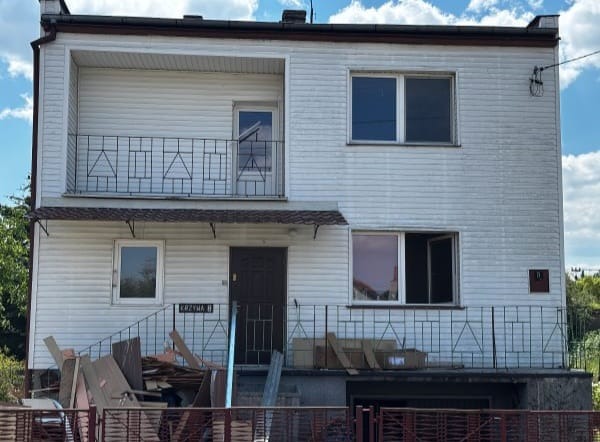 Dwupiętrowy dom z balkonem przed remontem. Widoczne materiały budowlane i śmieci przed wejściem. Elewacja z białego sidingu.