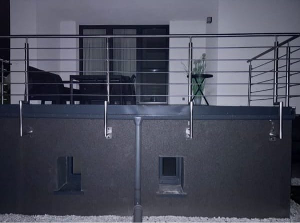 Nowoczesny balkon z metalową balustradą i ciemną elewacją. Widoczne małe okienka w dolnej części ściany i rynna. Na balkonie stolik z rośliną.