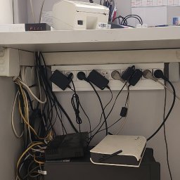 GLUVOLT Michał Głuchy - Nieuporządkowane kable i przedłużacz z wpiętymi zasilaczami pod blatem biurka. Widoczne routery i inne urządzenia elektroniczne. Chaos kabli.
