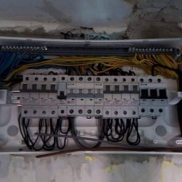 GLUVOLT Michał Głuchy - Rozdzielnica elektryczna w białej obudowie, z widocznymi przewodami i bezpiecznikami, zamontowana na surowej ścianie w trakcie prac instalacyjnych.
