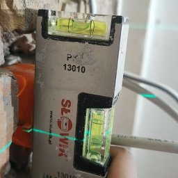 GLUVOLT Michał Głuchy - Poziomica laserowa w dłoni na tle surowej ściany z cegły i kabli. Precyzyjne wyznaczanie poziomu podczas prac instalacyjnych.