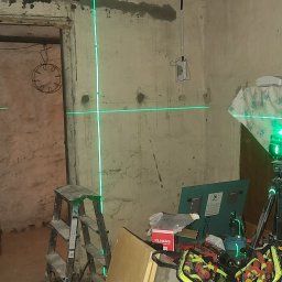 GLUVOLT Michał Głuchy - Laser krzyżowy wyznacza poziom na ścianach w trakcie remontu. Widoczna drabina, narzędzia i torba narzędziowa. Surowy stan ścian przed wykończeniem.