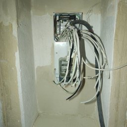 GLUVOLT Instalacje Elektryczne i Remonty - Otwarta skrzynka z licznikiem elektrycznym i wiązką kabli w surowym wnętrzu wnęki. Kable opisane markerem, ściany w trakcie wykończenia.