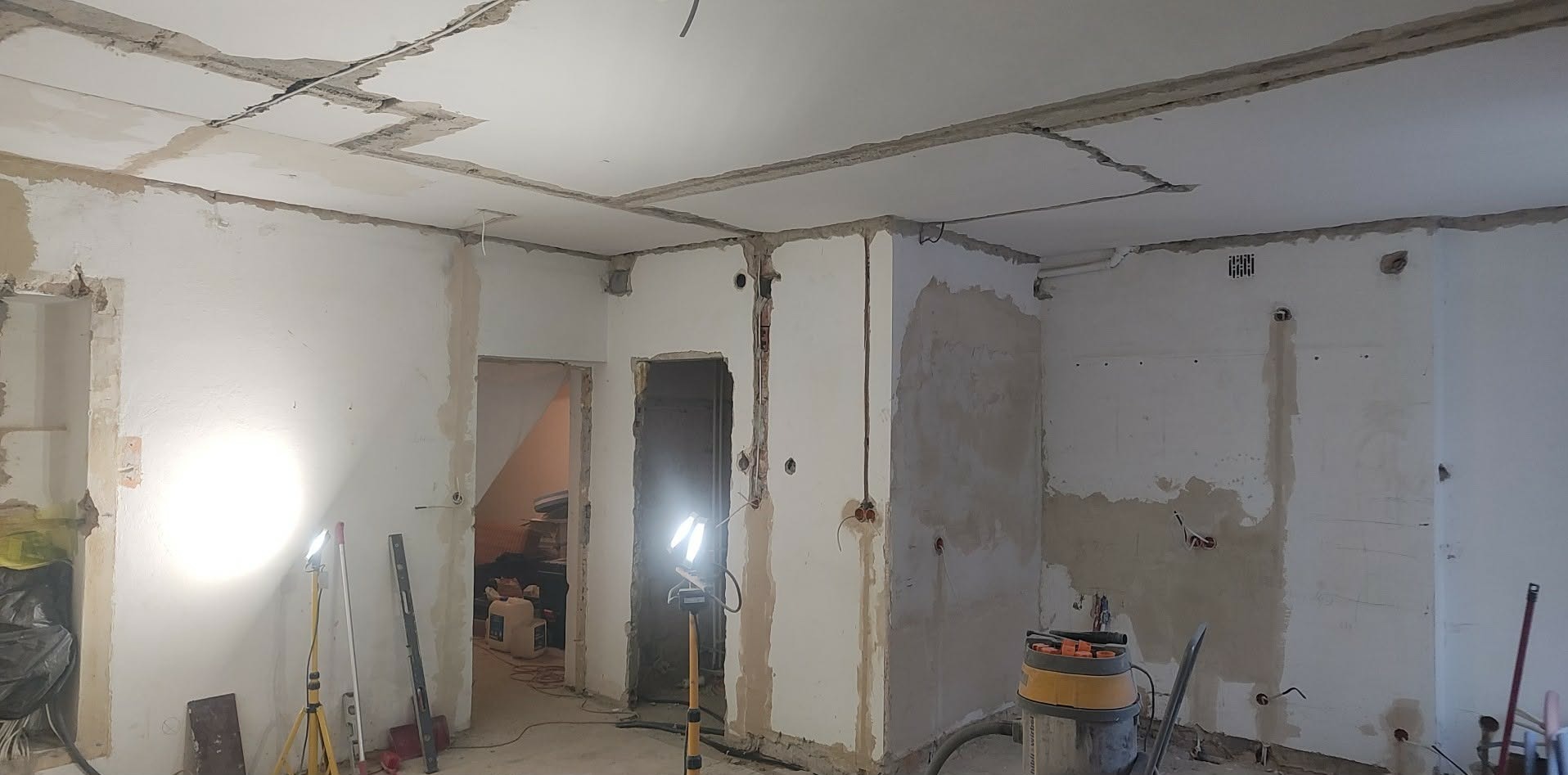 Wnętrze w trakcie remontu: odsłonięte przewody elektryczne, ściany przygotowane do tynkowania, widoczne narzędzia i lampy budowlane. Surowy stan przed wykończeniem.