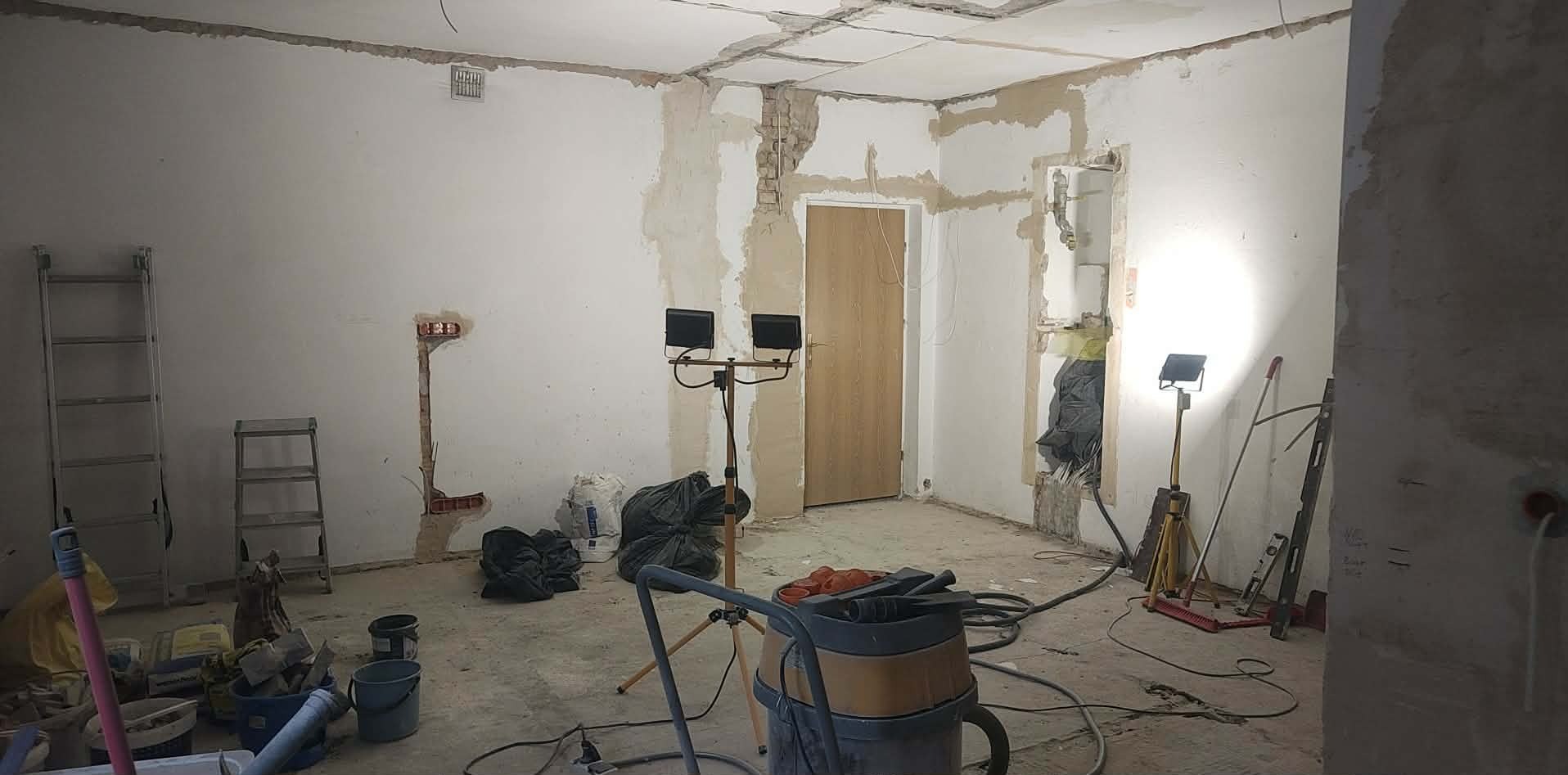 Wnętrze w trakcie remontu: odsłonięte fragmenty ścian, widoczne instalacje, narzędzia i worki z gruzem. Oświetlenie halogenowe na statywach. Etap przygotowania do wykończenia.