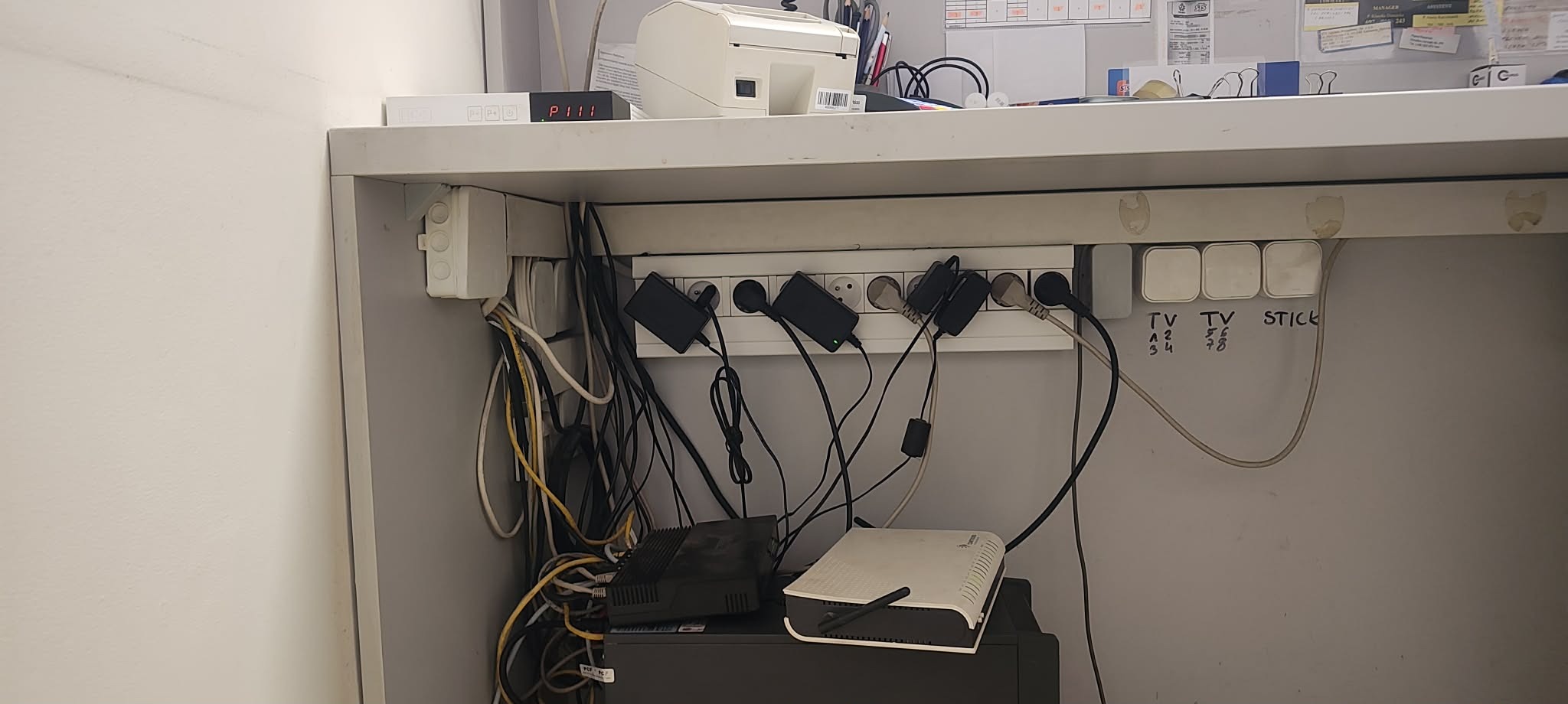 Nieuporządkowane kable i przedłużacz z wpiętymi zasilaczami pod blatem biurka. Widoczne routery i inne urządzenia elektroniczne. Chaos kabli.